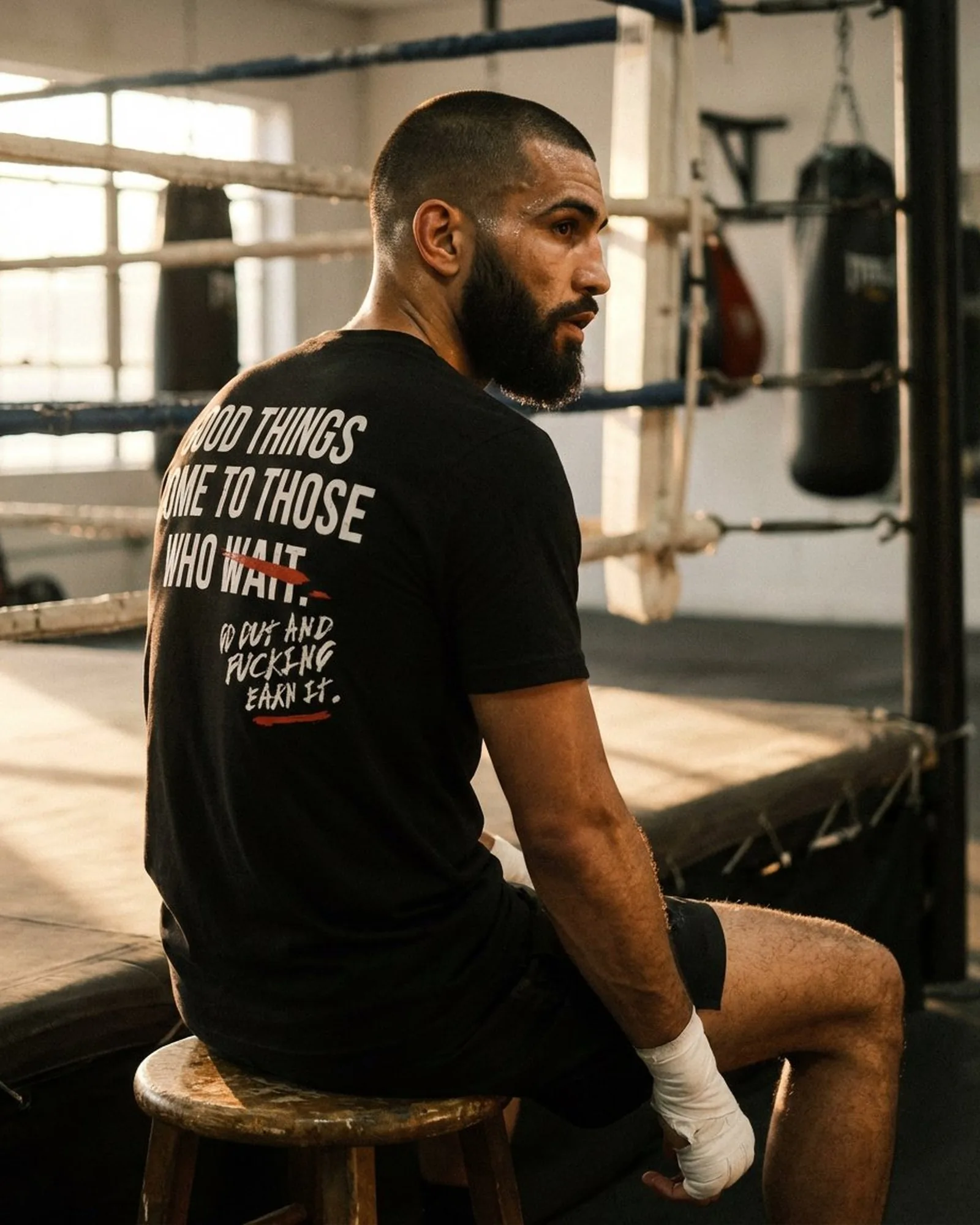 Wearing the Earn It tee in a boxing gym, sitting on a stool at the edge of the ring, full back graphic visible in warm window light
