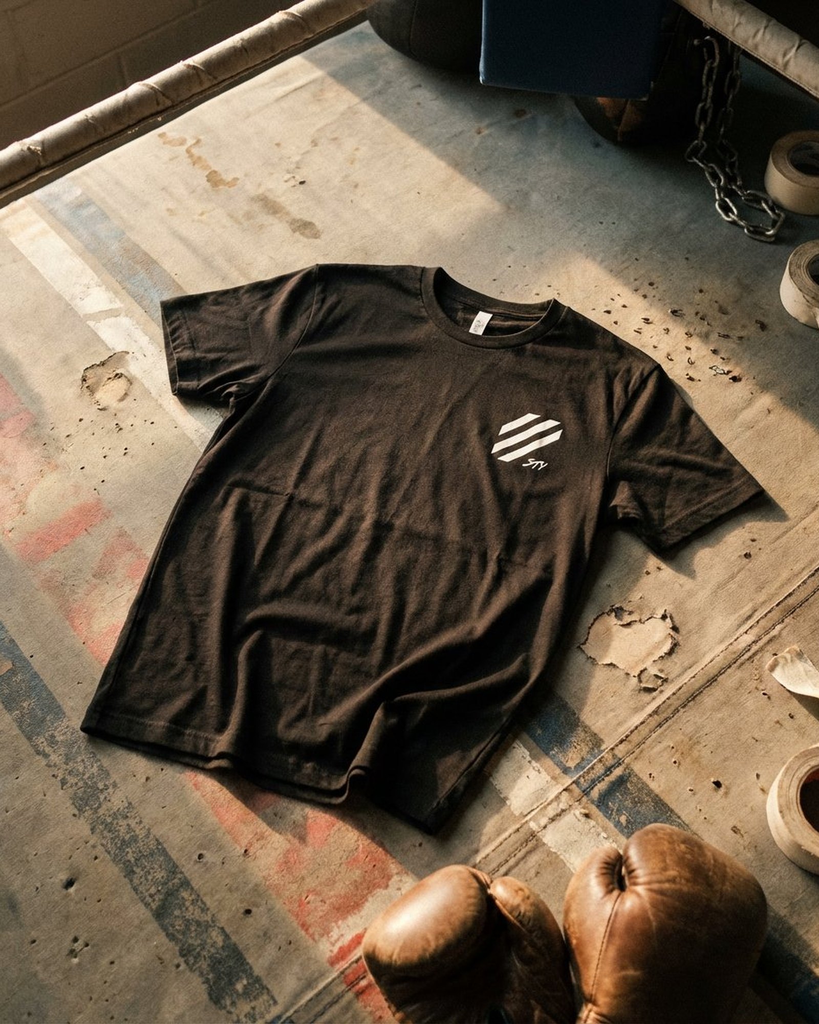 Earn It tee laid on weathered boxing-ring canvas in warm afternoon light, leather boxing gloves and hand-wrap tape visible at the edges, front STY hex-stripe mark on the chest
