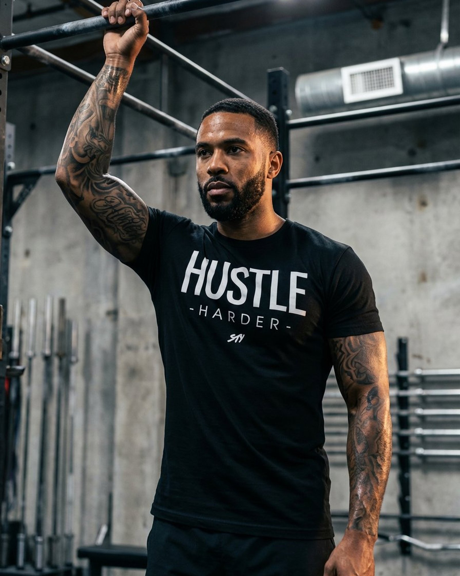 Wearing the Hustle Harder tee in the gym, hand on a pull-up bar, full tattoo sleeve visible, concrete and steel training environment