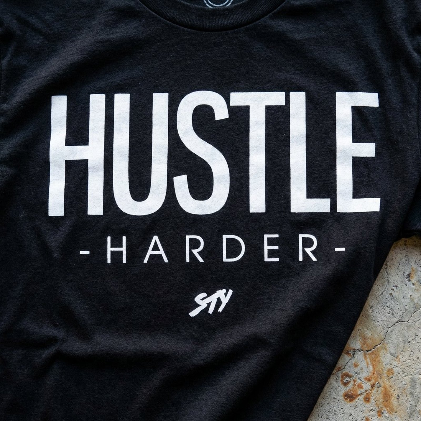 Macro closeup of the HUSTLE HARDER print on cotton fabric, weathered concrete edge visible at the corner