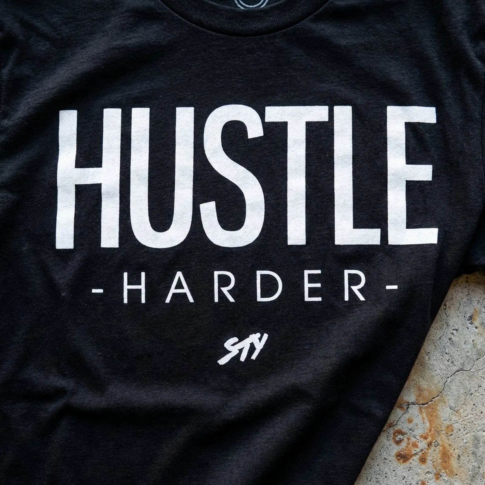 Macro closeup of the HUSTLE HARDER print on cotton fabric, weathered concrete edge visible at the corner